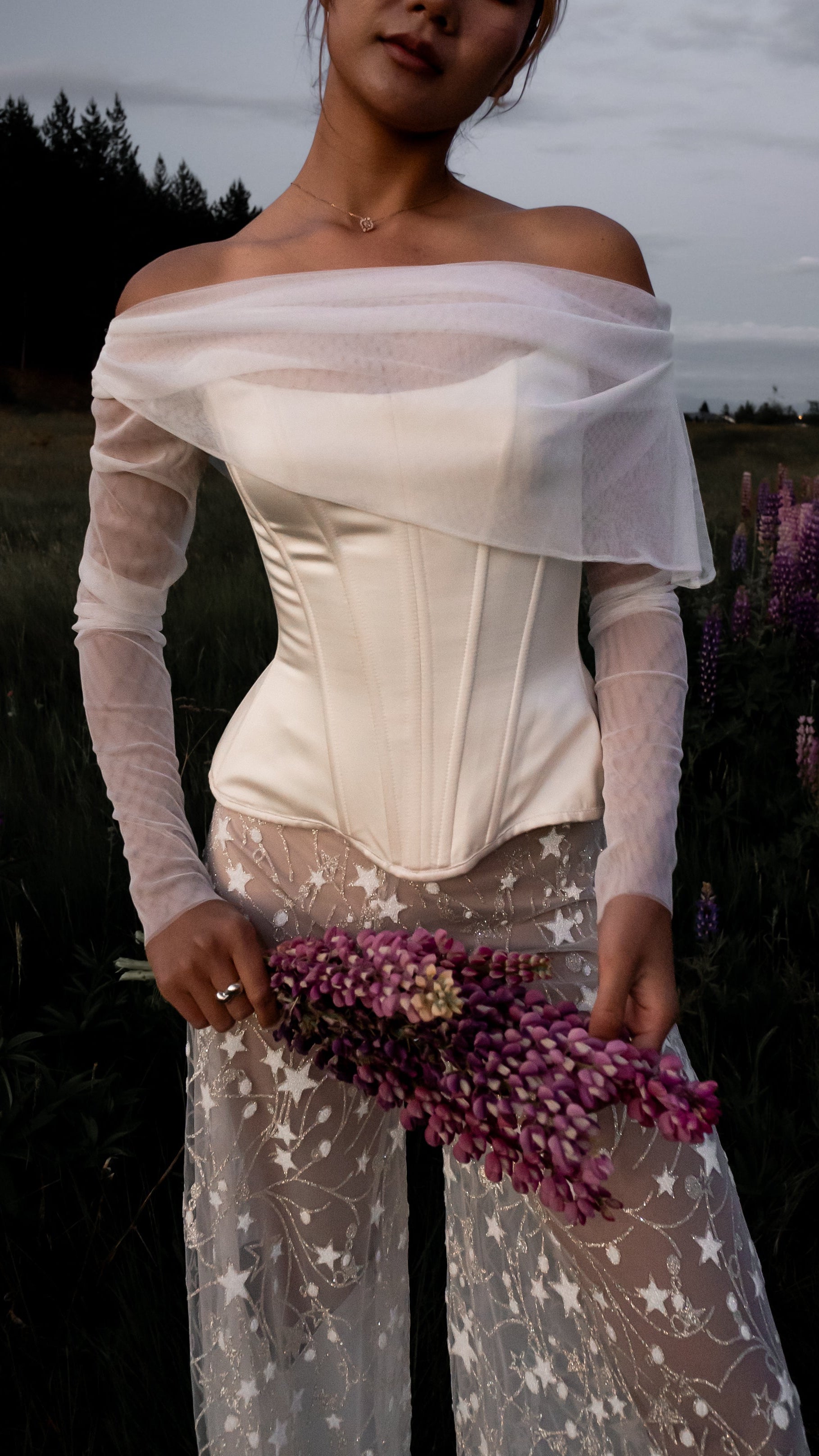 Bride in a white off shoulder long sleeve Basque waist corset dress with star pattern pants standing in a field of purple flowers.