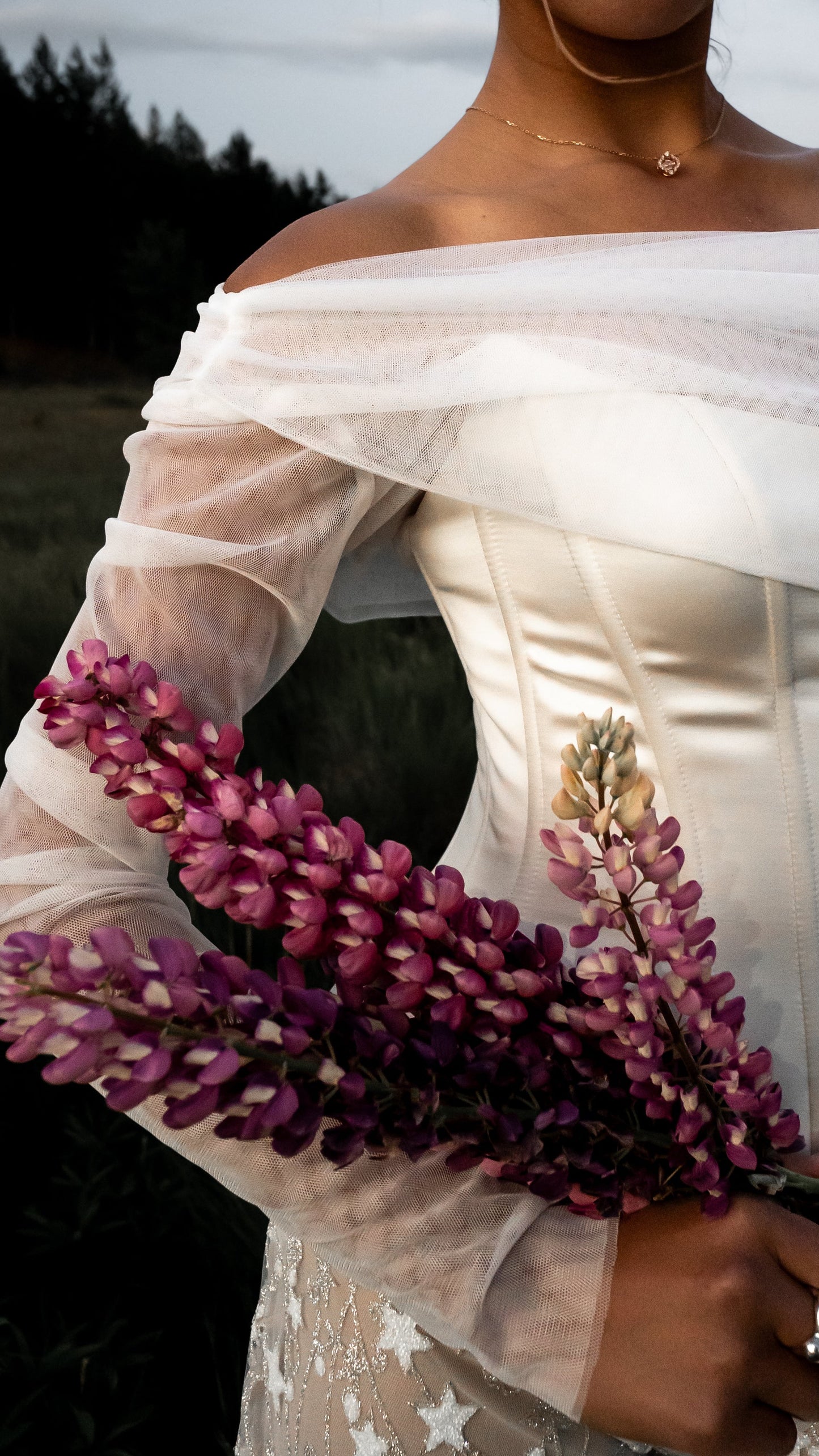 Bride in a white off shoulder Basque waist corset dress with star pattern pants standing in a field of purple flowers.