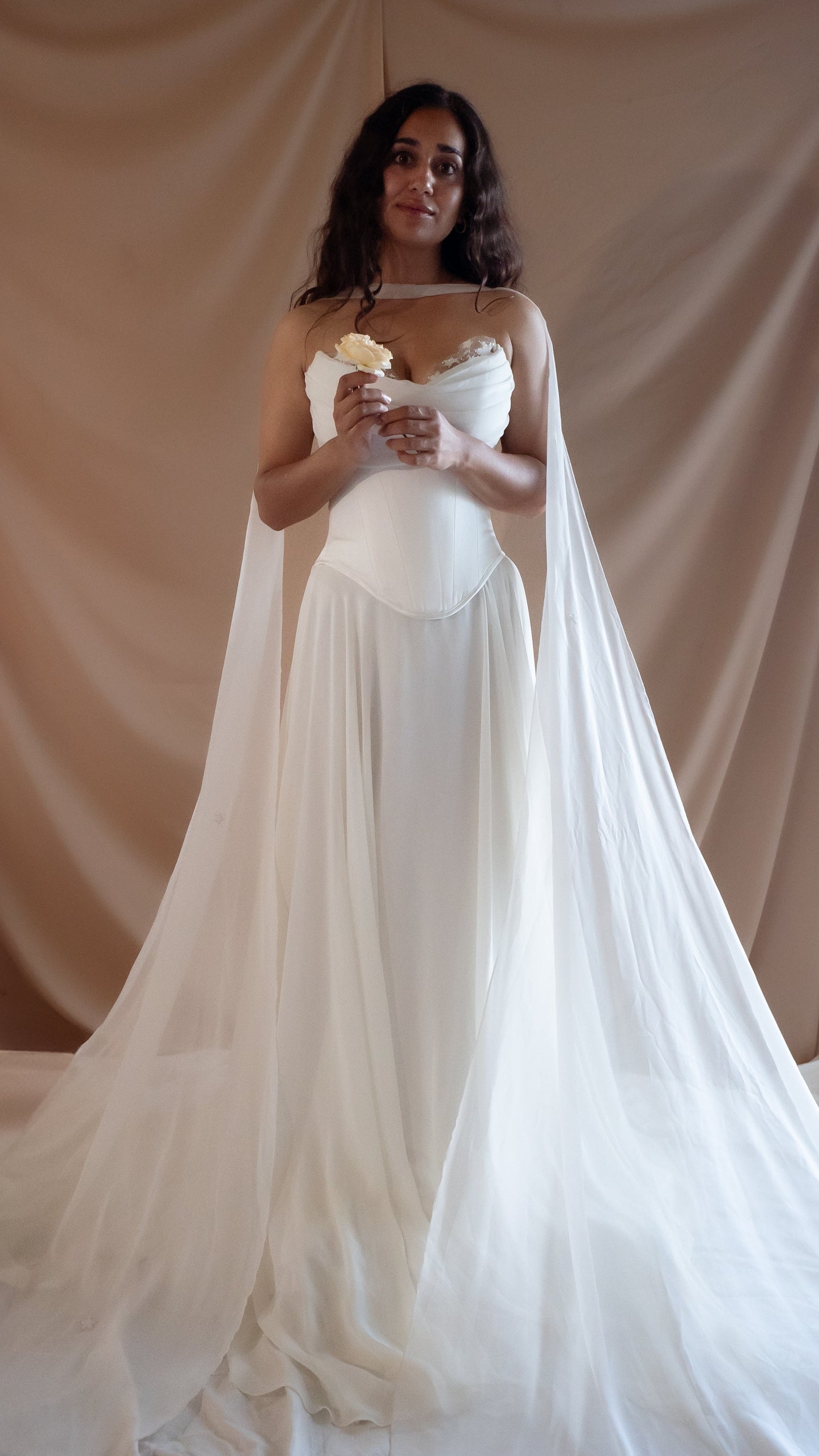 Woman in a vintage Basque waist wedding dress with a long train and neck chiffon scarf against a beige curtain backdrop