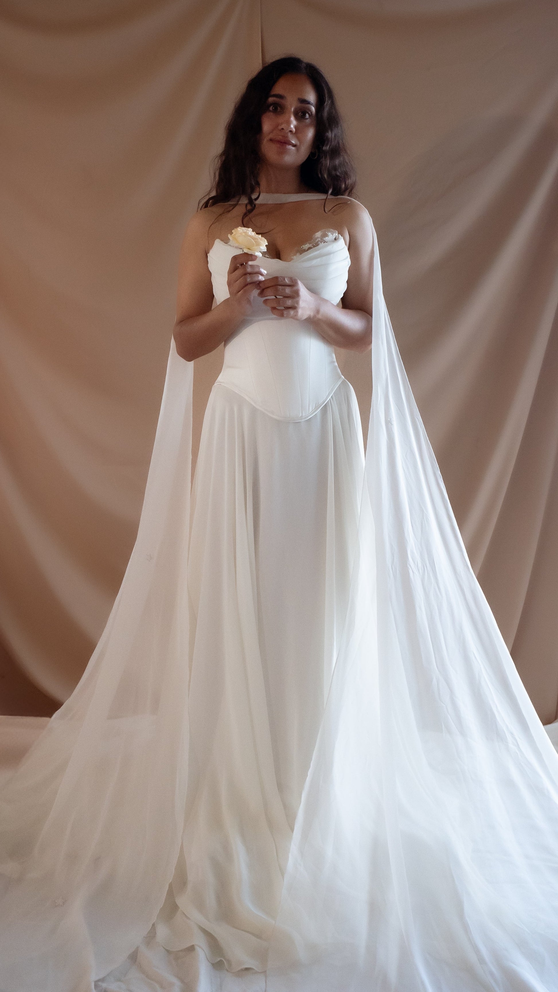 Woman in a vintage Basque waist wedding dress with a long train and neck chiffon scarf against a beige curtain backdrop